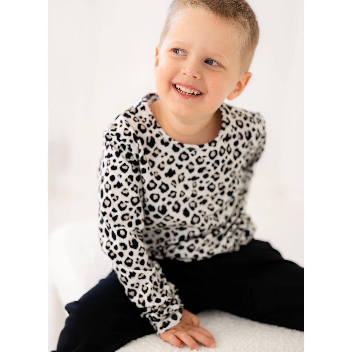 Poika istuu valkoisella penkillä, yllään mustavalkoinen leopardi printtipaita ja mustat housut, katsoo hymyillen sivulle