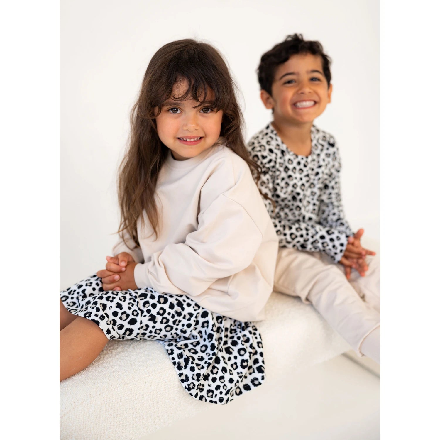 Lapset pukeutuneina Gugguun beigeihin ja leopardikuosisiin vaatteisiin, istuvat valkoisella penkillä