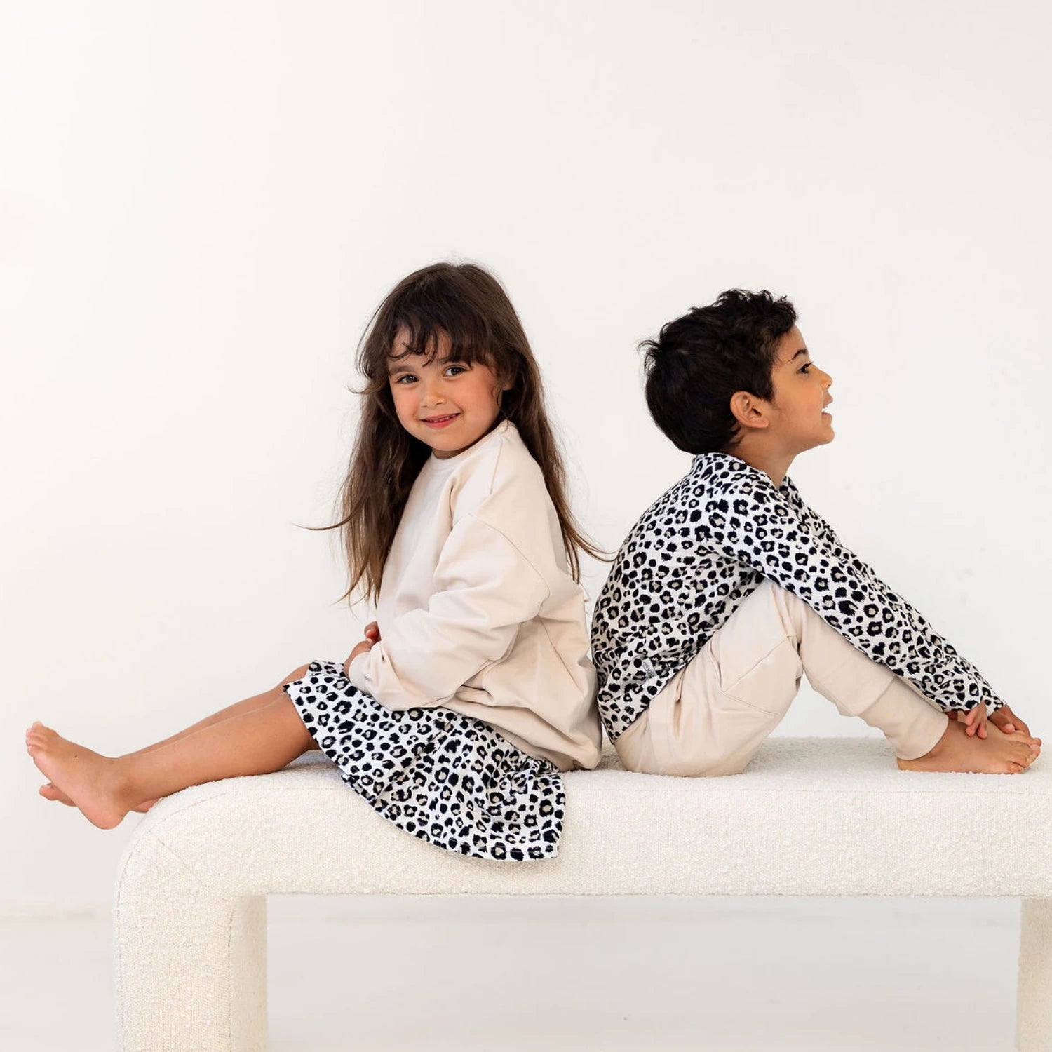 Lapset istuvat selät vastakkain, pukeutuneina Gugguun beigeihin ja leopardikuosisiin vaatteisiin