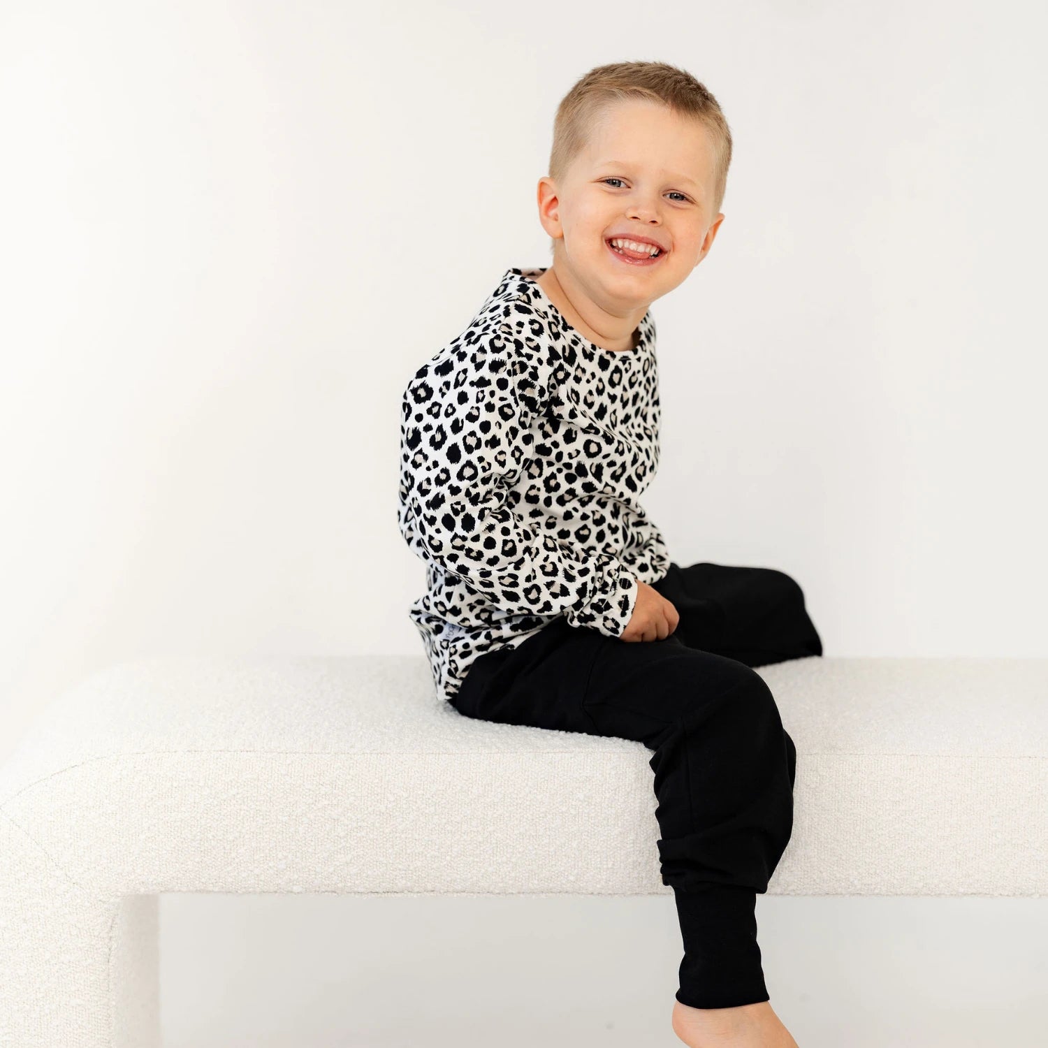 Poika istuu valkoisella penkillä, yllään rento leopardi printtipaita ja mustat housut, hymyilee kameralle