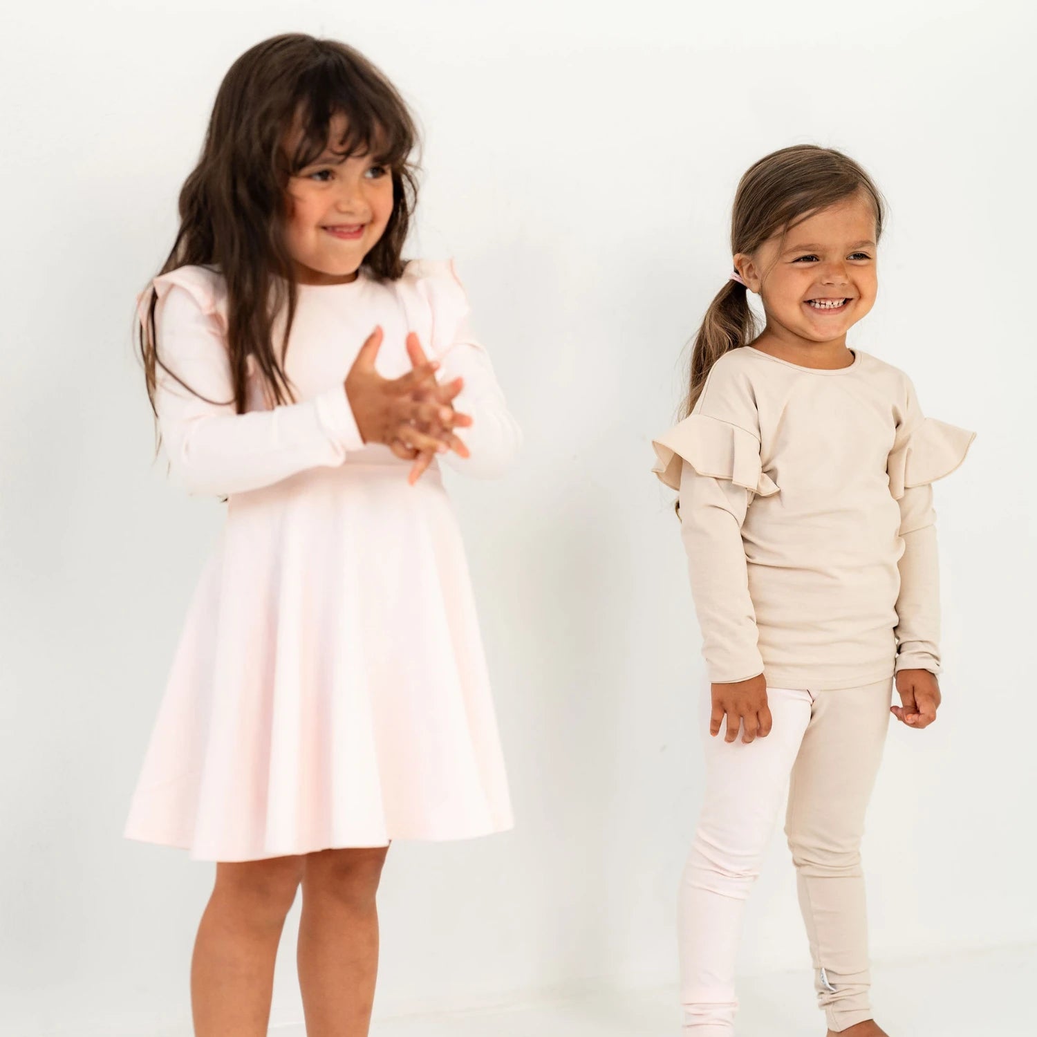 Kaksi tyttöä vaaleissa frillahihallisissa asuissa nauravaisina valkoista taustaa vasten