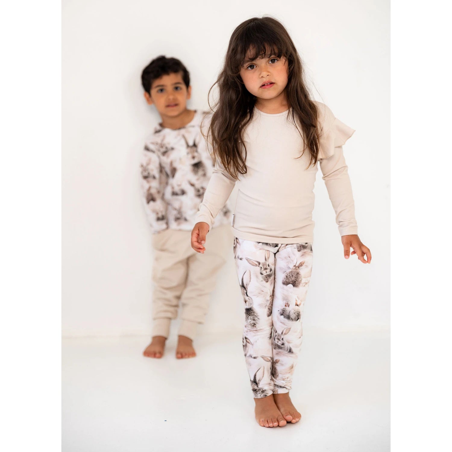 Kaksi lasta poseeraa yhdessä. Edessä lapsi pupu printtisissä leggingseissä ja yksivärisessä paidassa ja taustalla lapsi saman sarjan collegepaidassa ja beigessä alaosassa.
