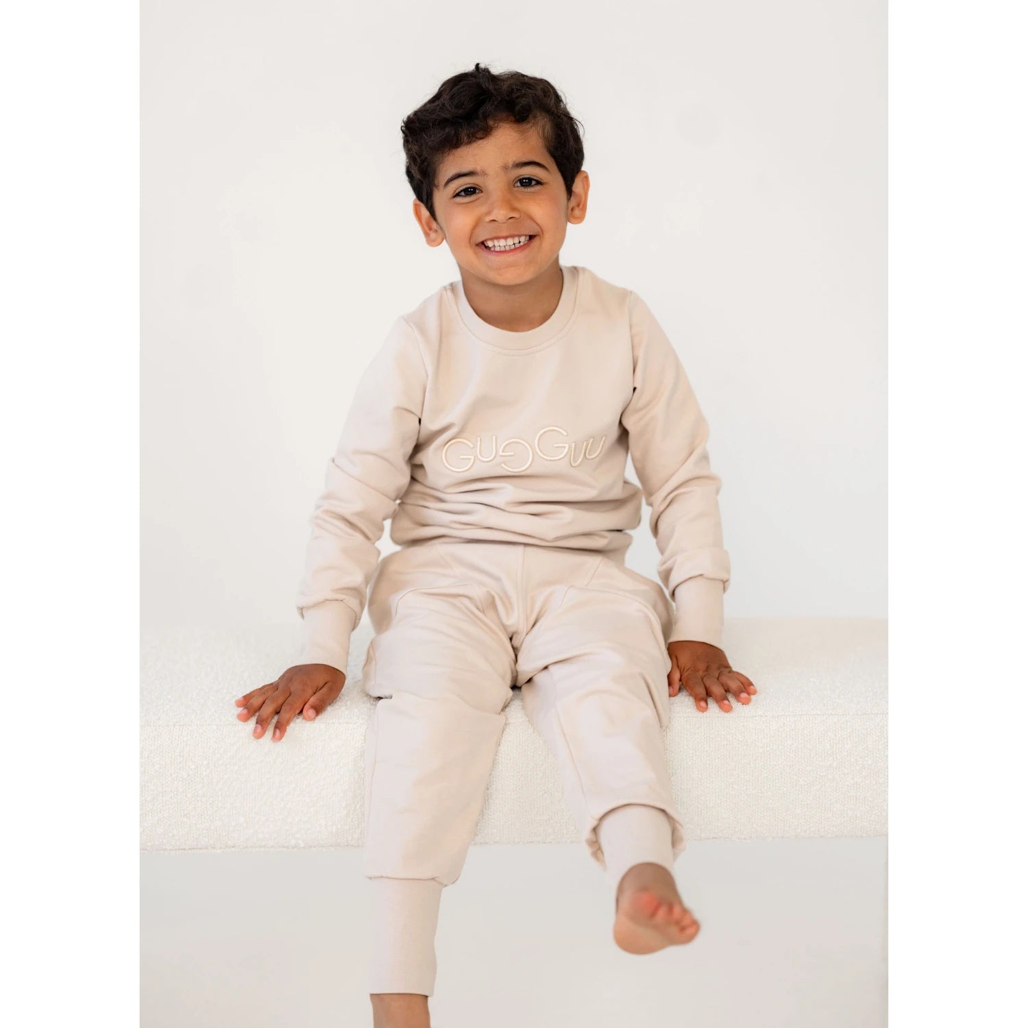 Poika beigessä collegesetissä istuu valkoisella penkillä ja hymyilee iloisesti kameraan




