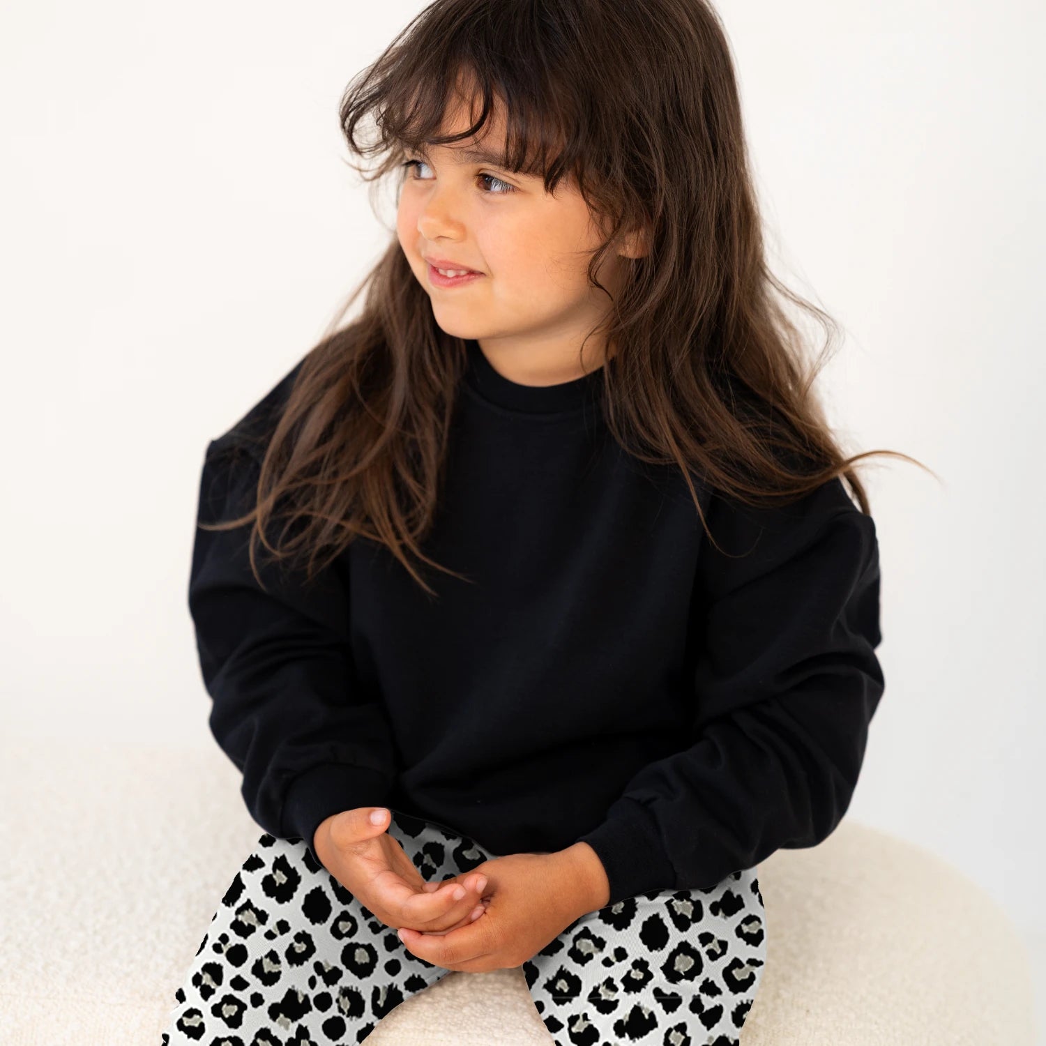 Tyttö istuu vaalealla penkillä, pukeutuneena mustaan collegepaitaan ja leopardikuosisiin leggingseihin