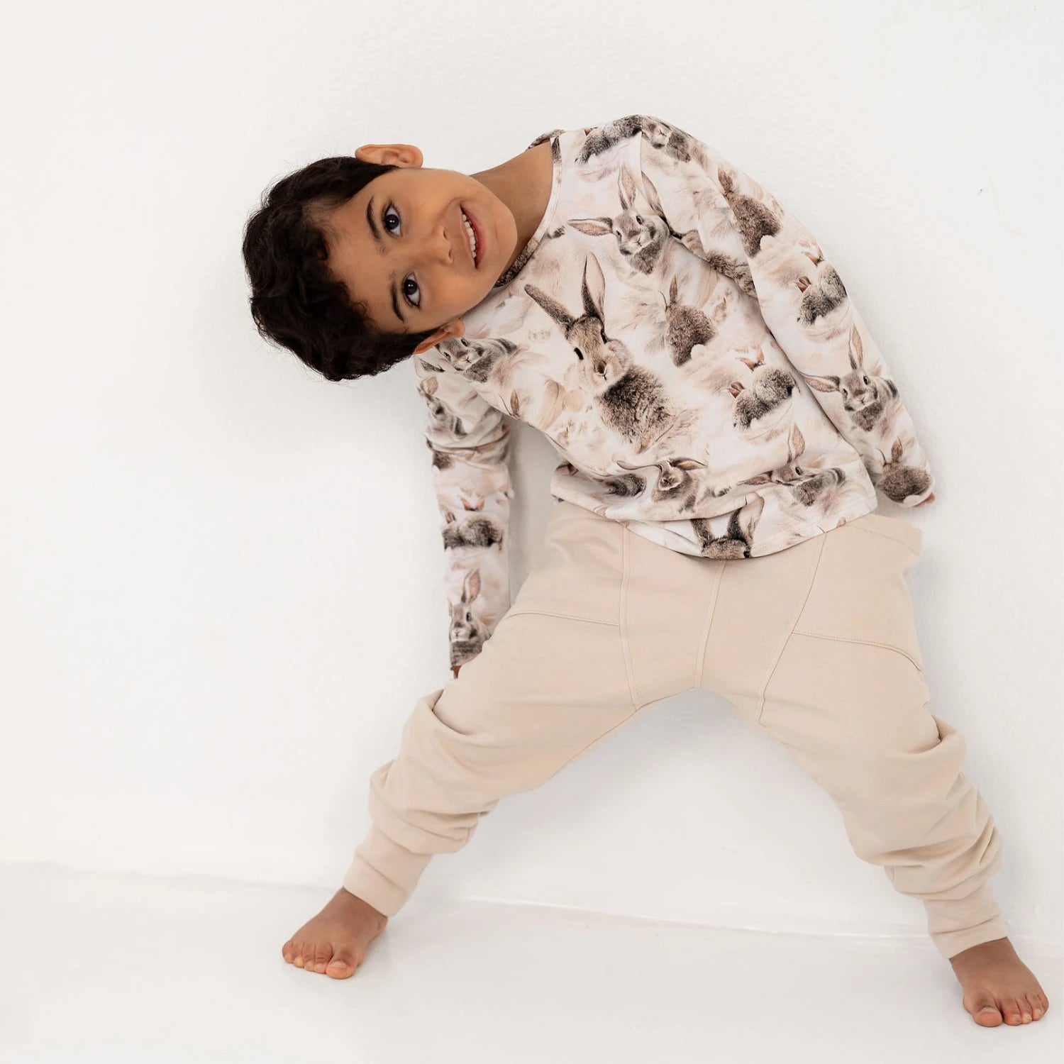 Poika nojaa seinää vasten pupun printtisessä pitkähihaisessa ja beigeissä housuissa