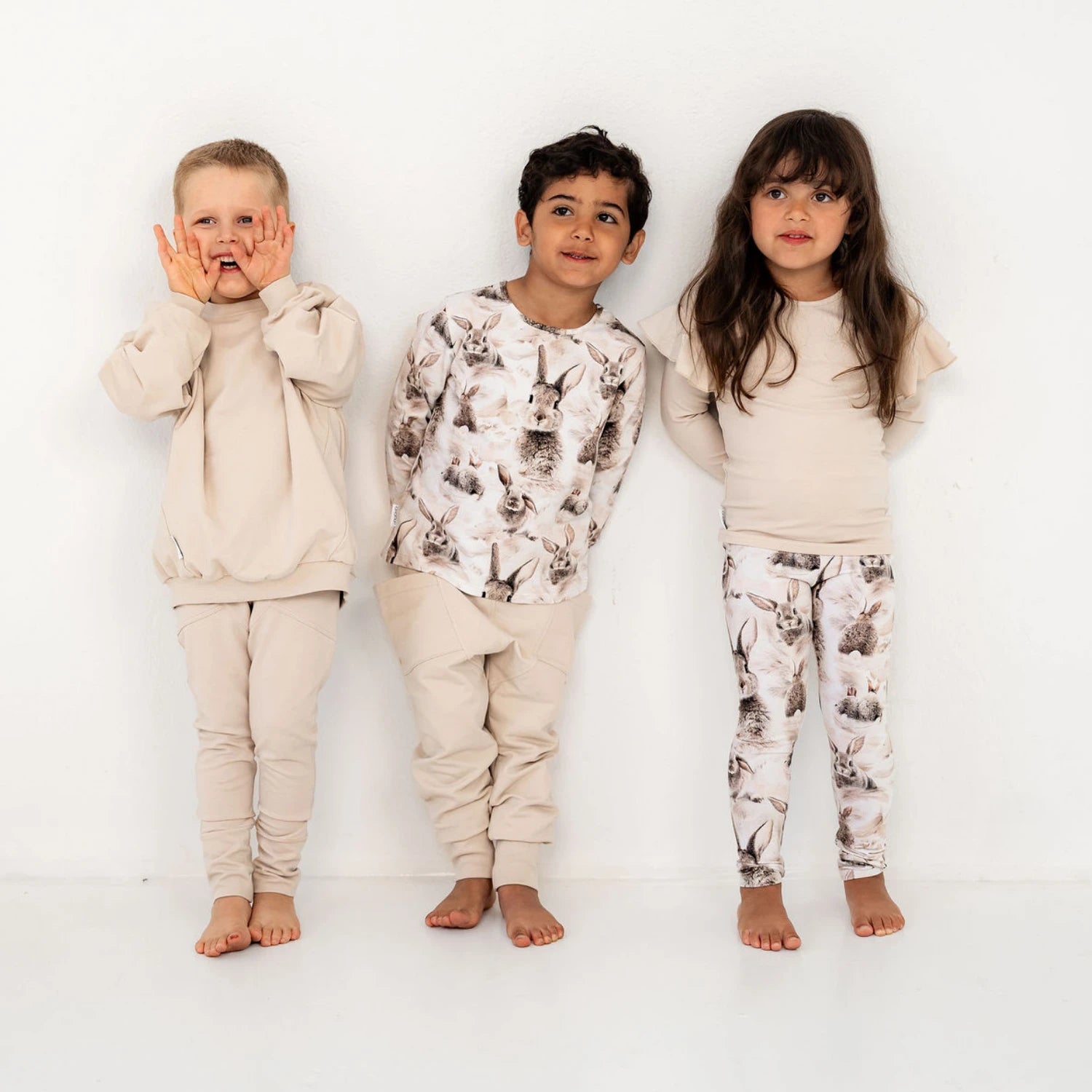 Kolme lasta seisoo vierekkäin. Keskellä olevalla pupu printtinen paita ja beiget housut, oikealla pupu legginsit ja frillapaita sekä vasemmalla olevalla beige asukokonaisuus