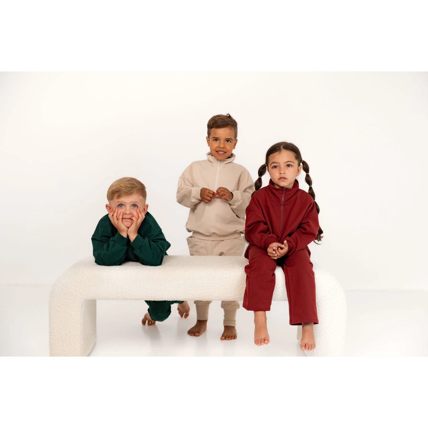 Kolme lasta oleskelemassa penkillä. Kaikkilla on päällä puoli vetoketjullinen collegepaita ja collegehousut