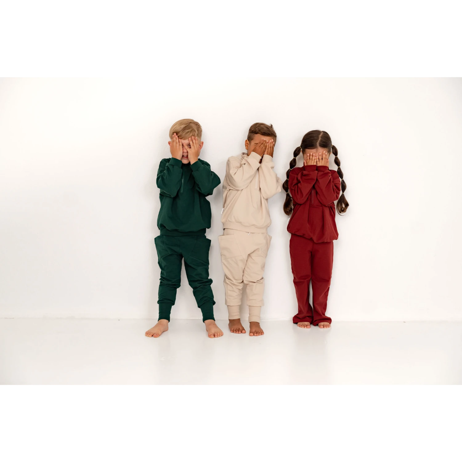 [ALL] Kolme leikkisää lasta seinää vasten kädet naaman edessä. Lapsilla on suloiset collegesetit päällä