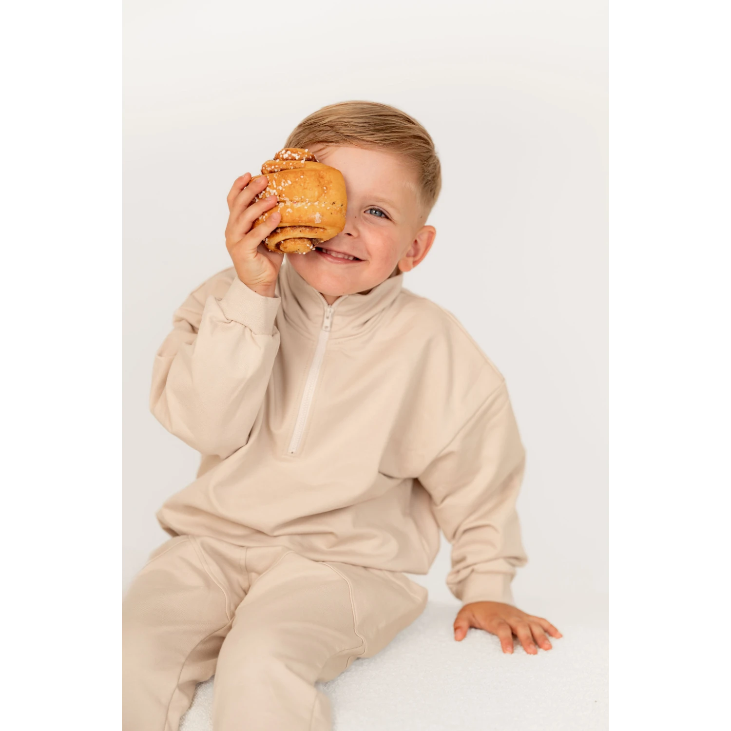 Poika hymyillee kameralle pulla kädessä. Lapsell aon päällä rento collegesetti tyylikkäillä yksityiskohdilla.