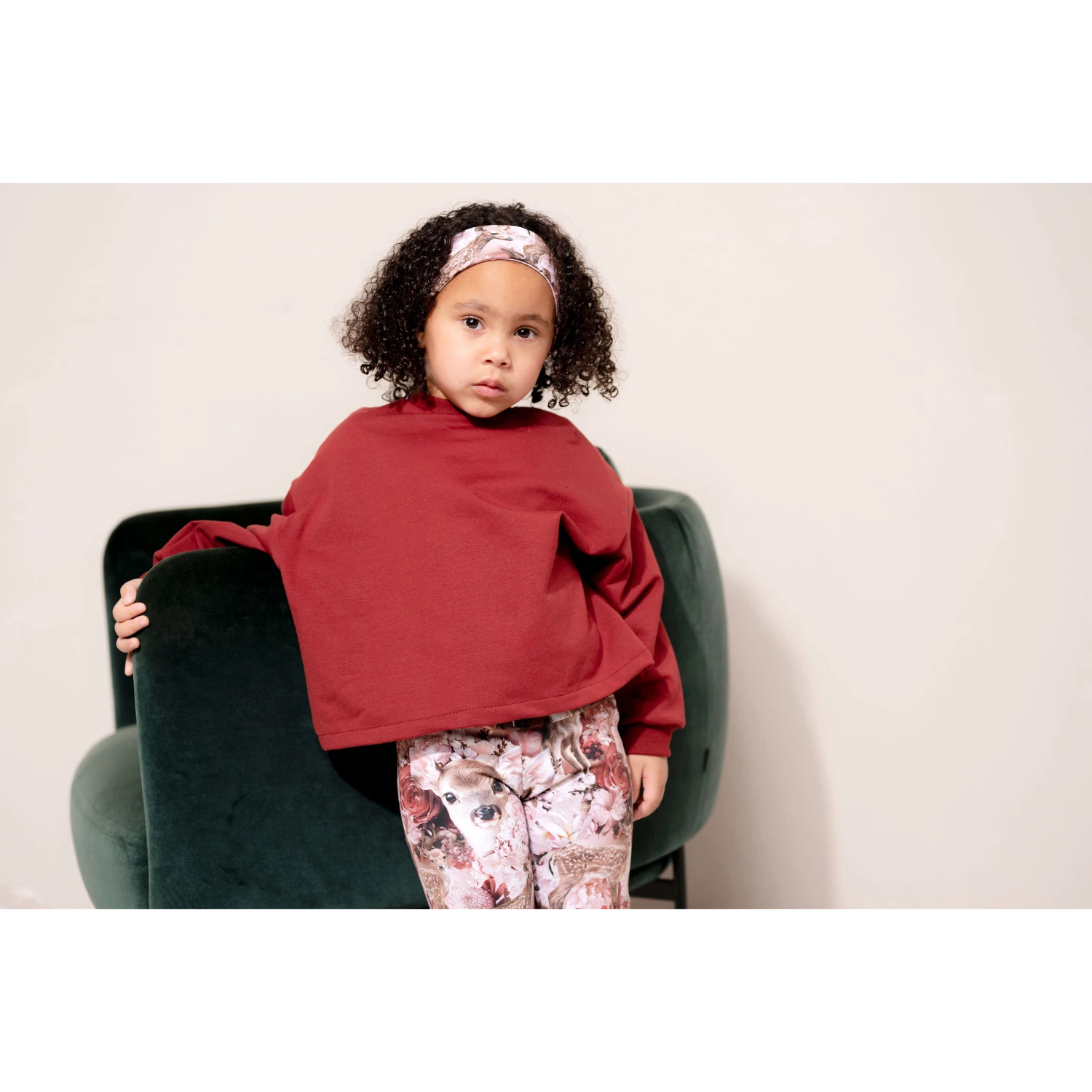 Tyttö nojaa sohvaa vasten ja katsoo kameraan. Lapsella on päällä tummanpunainen collegepaita ja bambi kuosilliset leggingsit